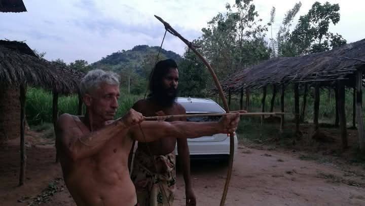 Solo traveler practicing archery with a wooden bow in an outdoor rural setting with thatched structures.